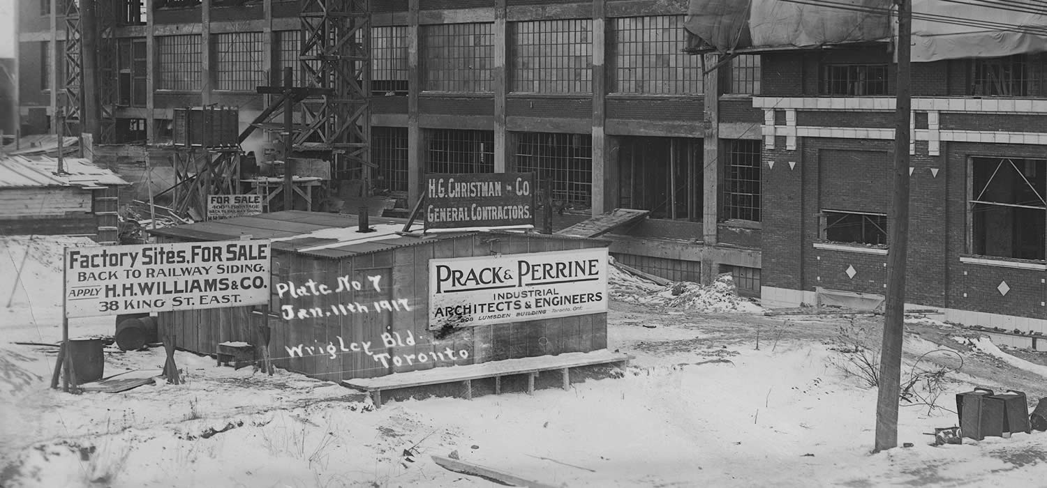 A factory building under construction under construction in winter. The upper floors are a concrete skeleton while the lower floors are approaching completion. Signs advertising the architect, general contractor, and real estate agent are next to the construction site.