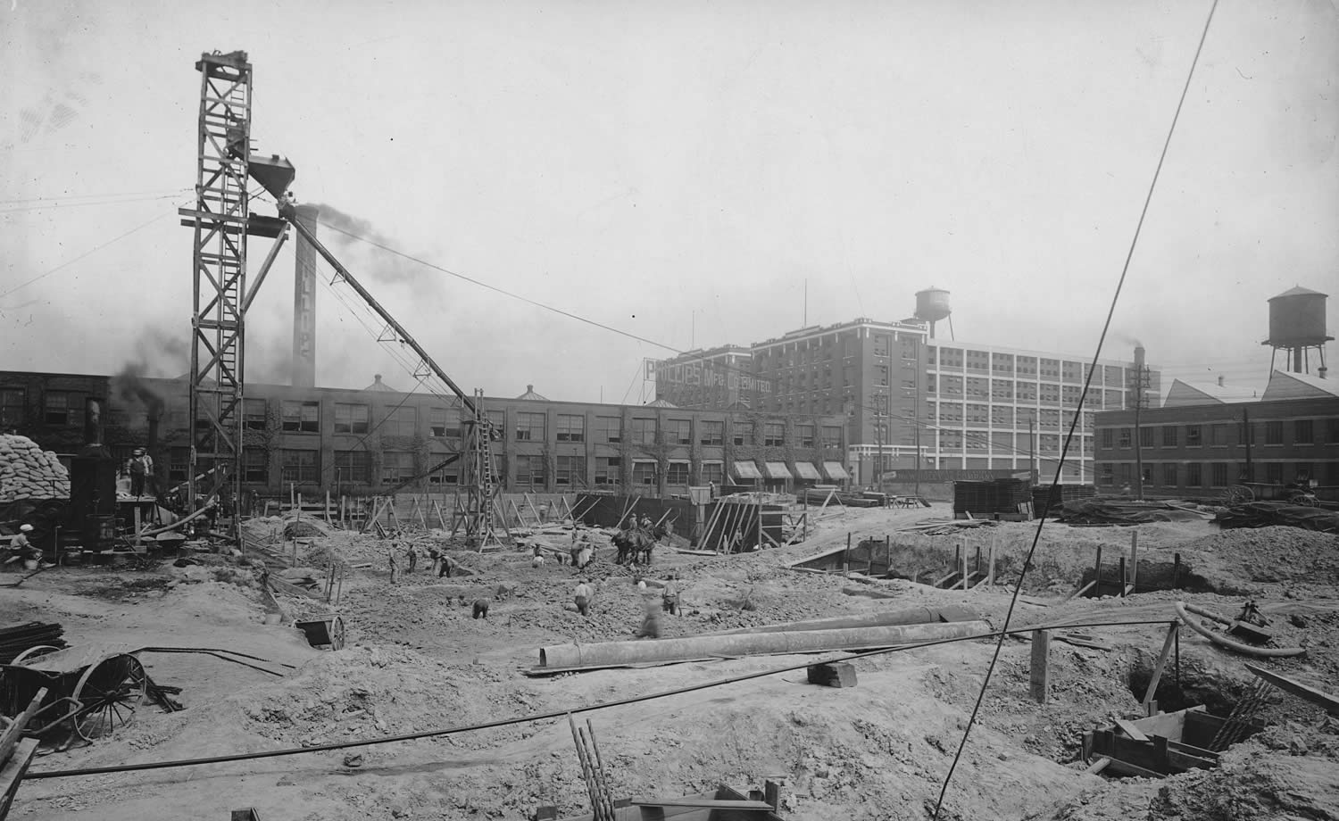 Workers with horses laying the foundation for a factory building with other completed factories in the background.