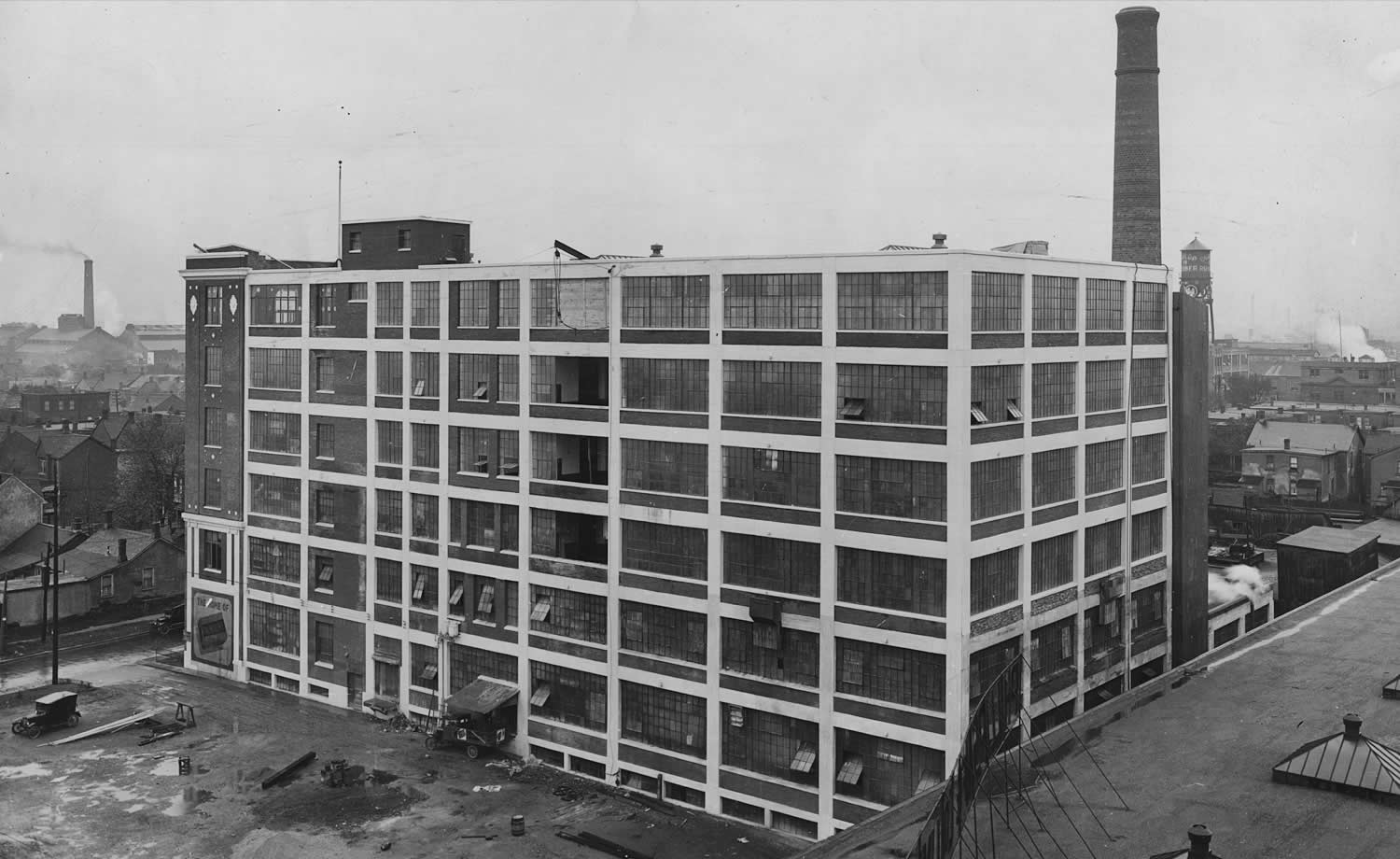 Black and white photograph of a large brick factory building with a chimney stack at the back. Large windows.