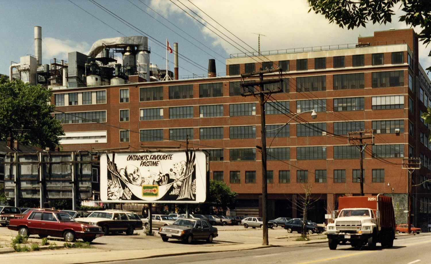Colour photograph of the Colgate-Palmolive factory on Carlaw Avenue showing almost the entirety of the south facade. A parking lot containing a billboard advertising the Lottario lottery is in the foreground.