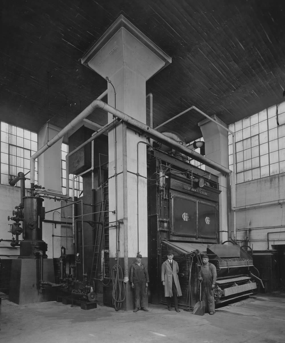 Two workers and a man in smart clothing in front of a machine, likely a soap kettle, inside the Palmolive Co. of Canada factory on Natalie Avenue.