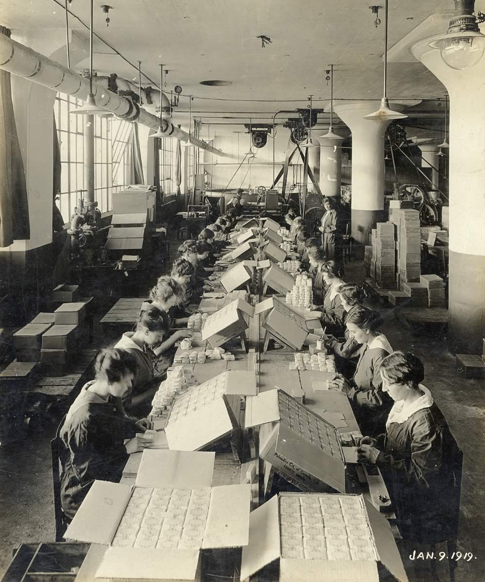 Sepia photograph of a group of women of various ages on a production line wrapping and packing soap by hand. Unwrapped bars are on a conveyor belt in the middle of their table and the women are wrapping them in paper and putting them in cardboard boxes. A female supervisor is visible in the background.
