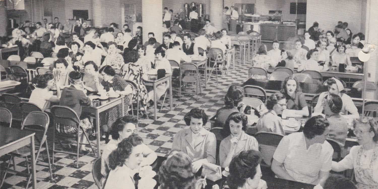 Black and white photograph taken of a busy cafeteria full of men and women eating and talking. Checkered flooring.