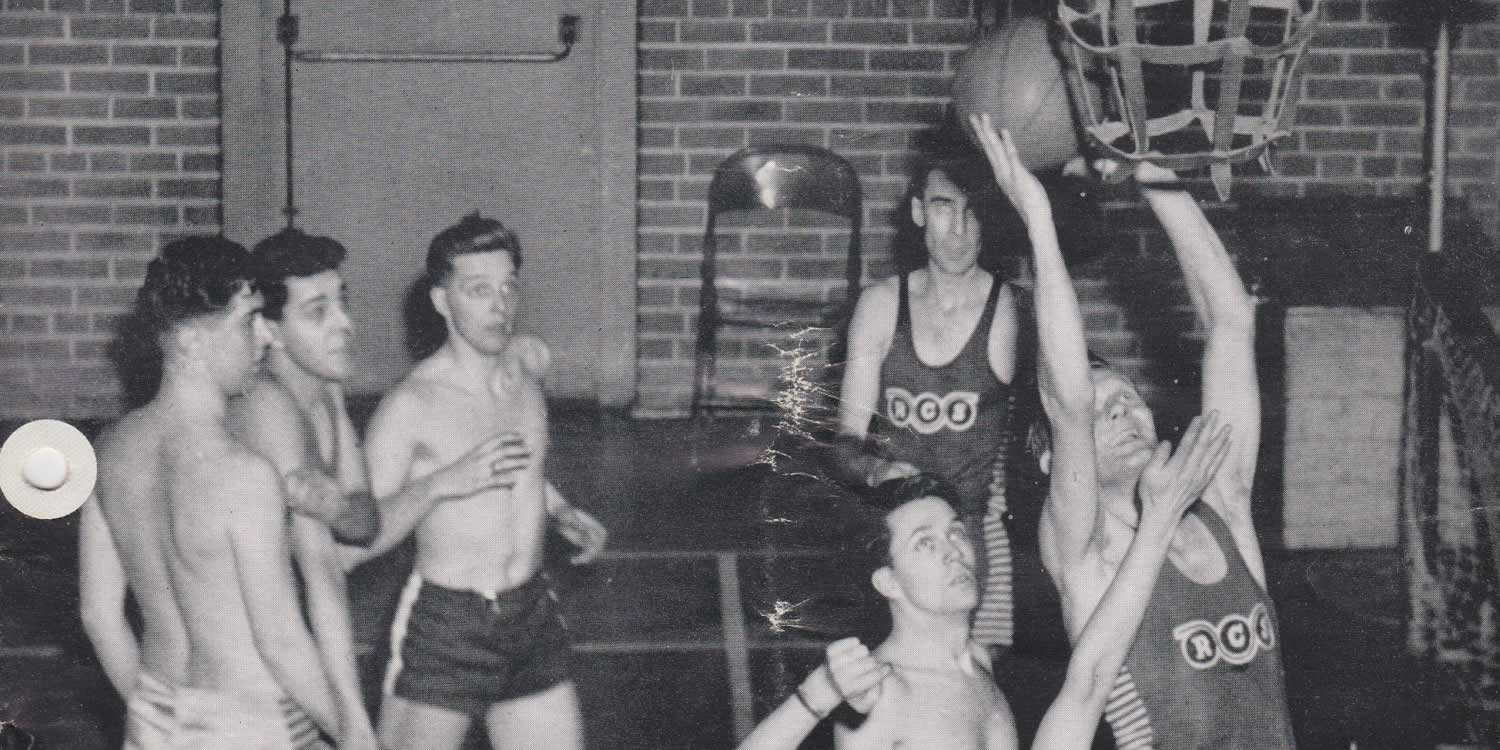 Black and white photograph taken on a basketball court. Seven Men in 1950s basketball gears are vying in front of the basket, some are shirtless, others have 'RCS' on their jerseys.