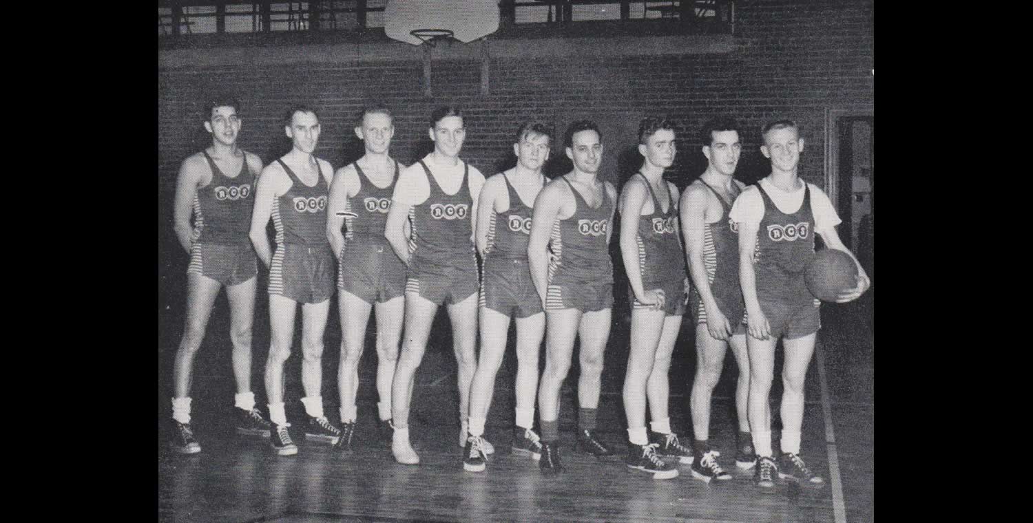 Black and white photograph of a basketball team wearing 1950s shorts and jerseys, and high tops. The letters 'RCS' are written on their jerseys.