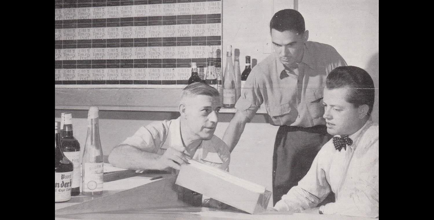 Black and white image of three men discussing a product package in an office. The men are dressed in shirts and one is wearing a bow tie.