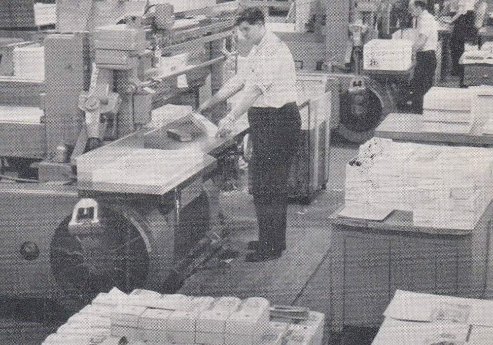 Black and white image of two men working on a printing press line. They are cutting printed papers. Stacks of printed certificates surround them.