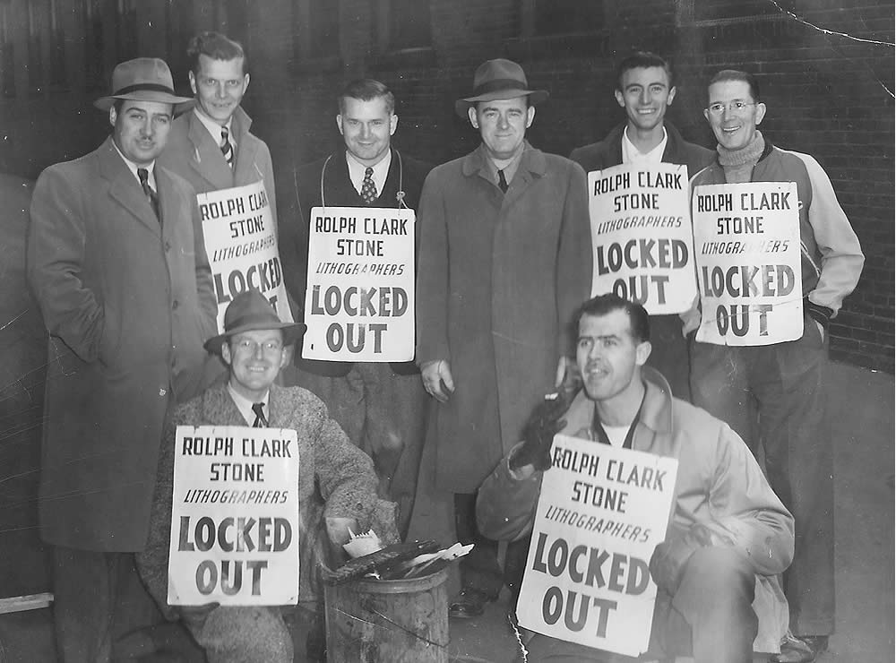 Black and white image of a group of nine men on a picket line holding 'locked out' placards. Two men are crouched in front. Many are wearing winter coats and hats.