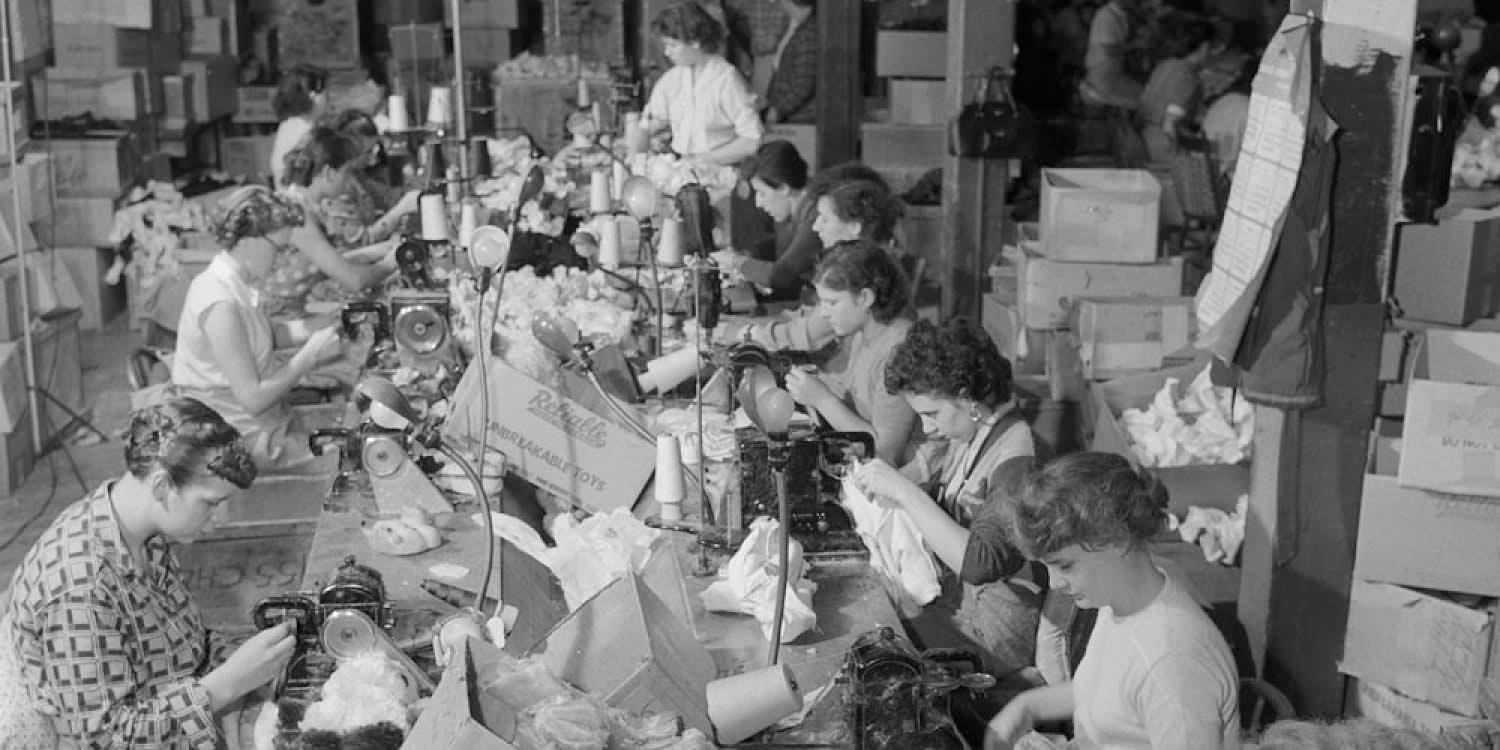 Archival image of a portion of an assembly line. A dozen women are seated at a long table sewing plush toys with sewing machines.