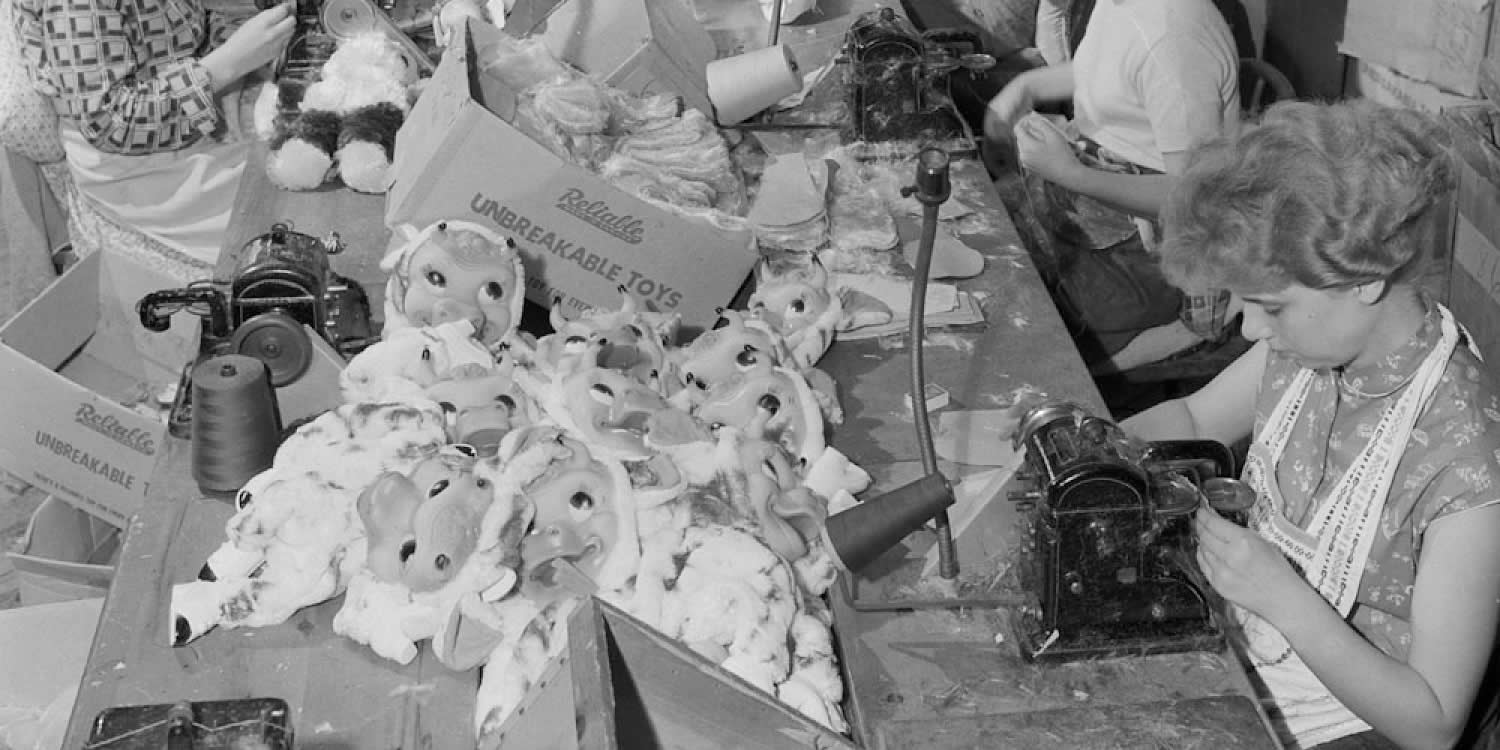 Archival image of a portion of an assembly line. A dozen women are seated at a long table sewing plush toys with sewing machines.