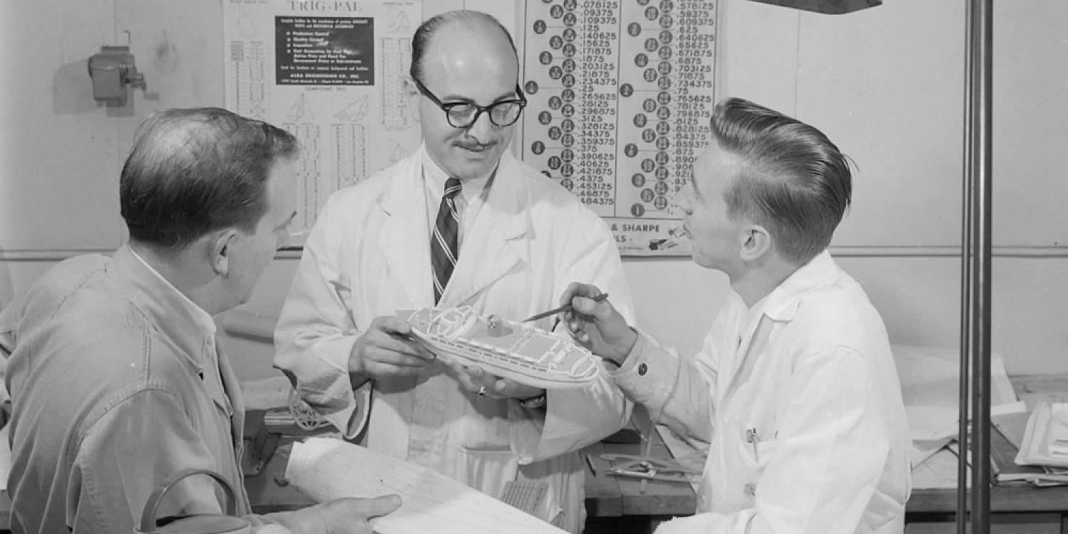 Black and white image of three men looking at small toy boat. Two men are wearing labcoats. Other toys are visible on the table.