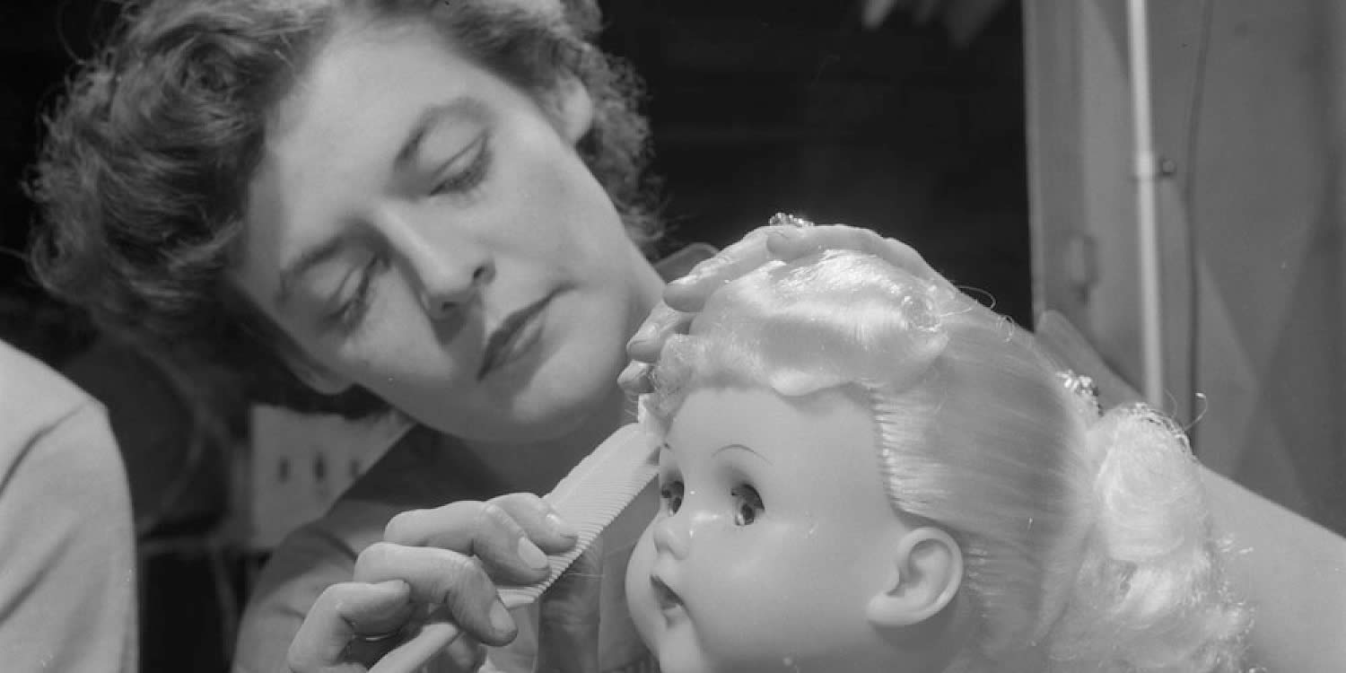 Black and white image of woman combing a doll's hair on a head that propped up on a piece of wood.