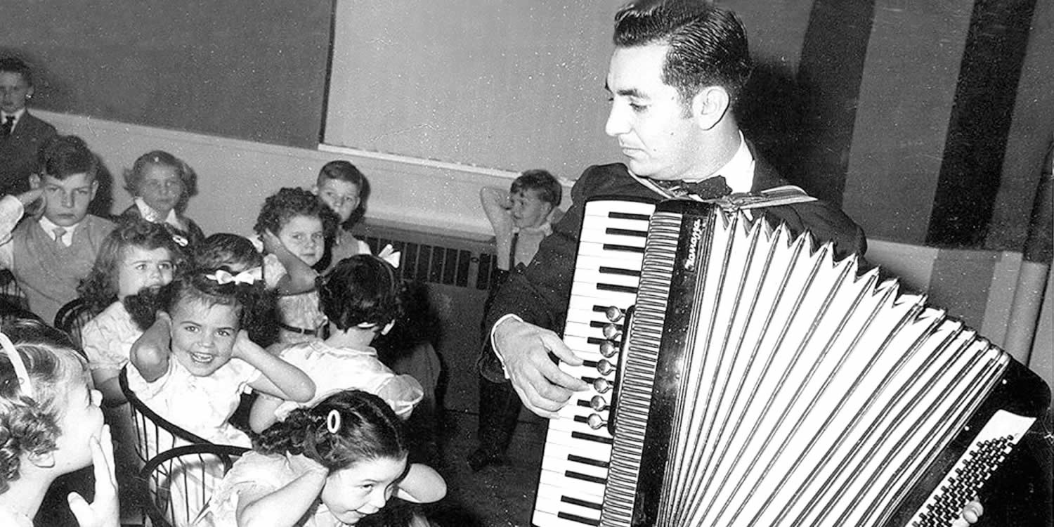 Black and white photograph of a man playing the accordion to a seated group of children. Some are covering their ears.