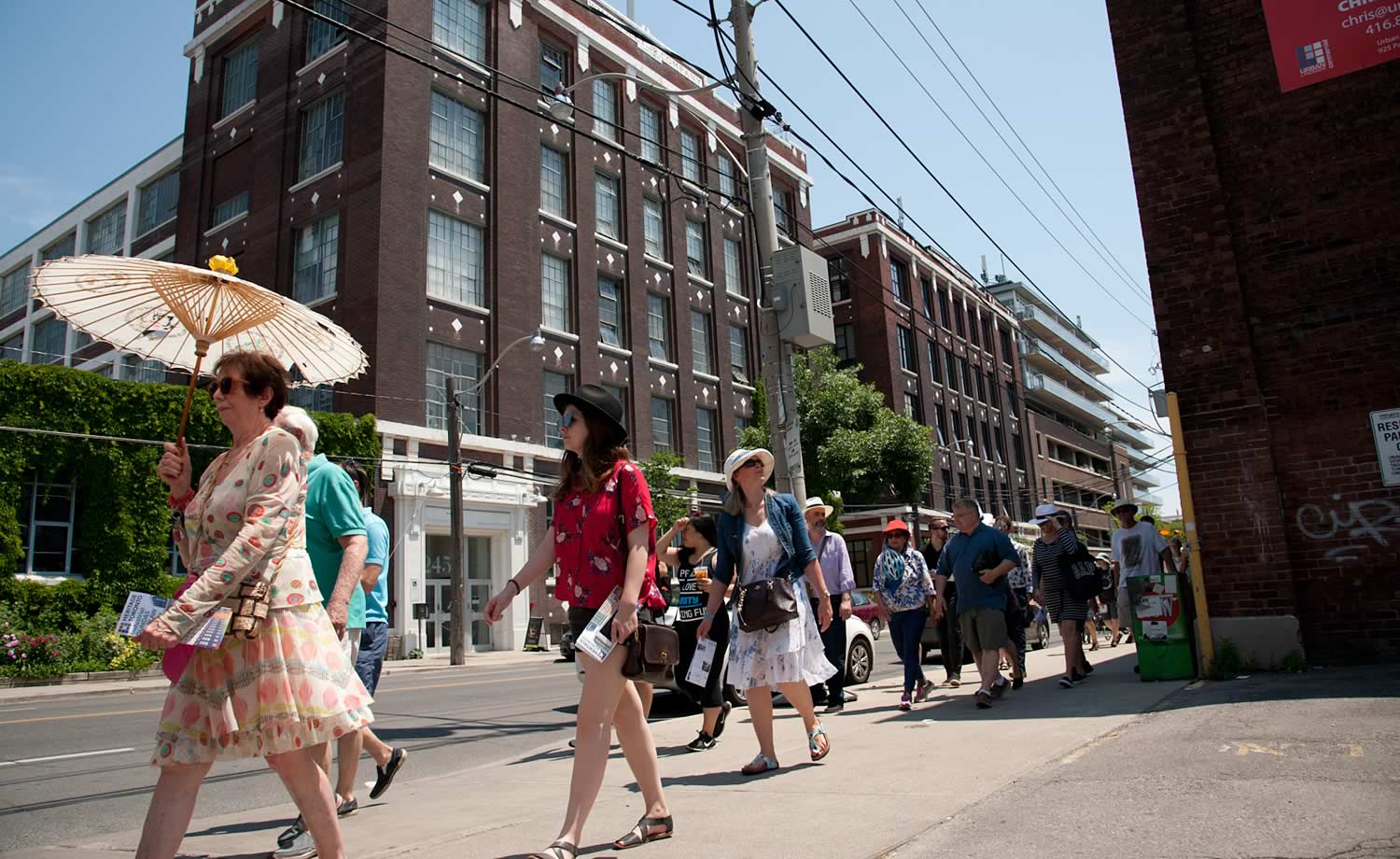Colour photograph of an industrial brick building divided into to aisles. A tour group walks past the building, on the other side of the street. A women is holding a sun umbrella.