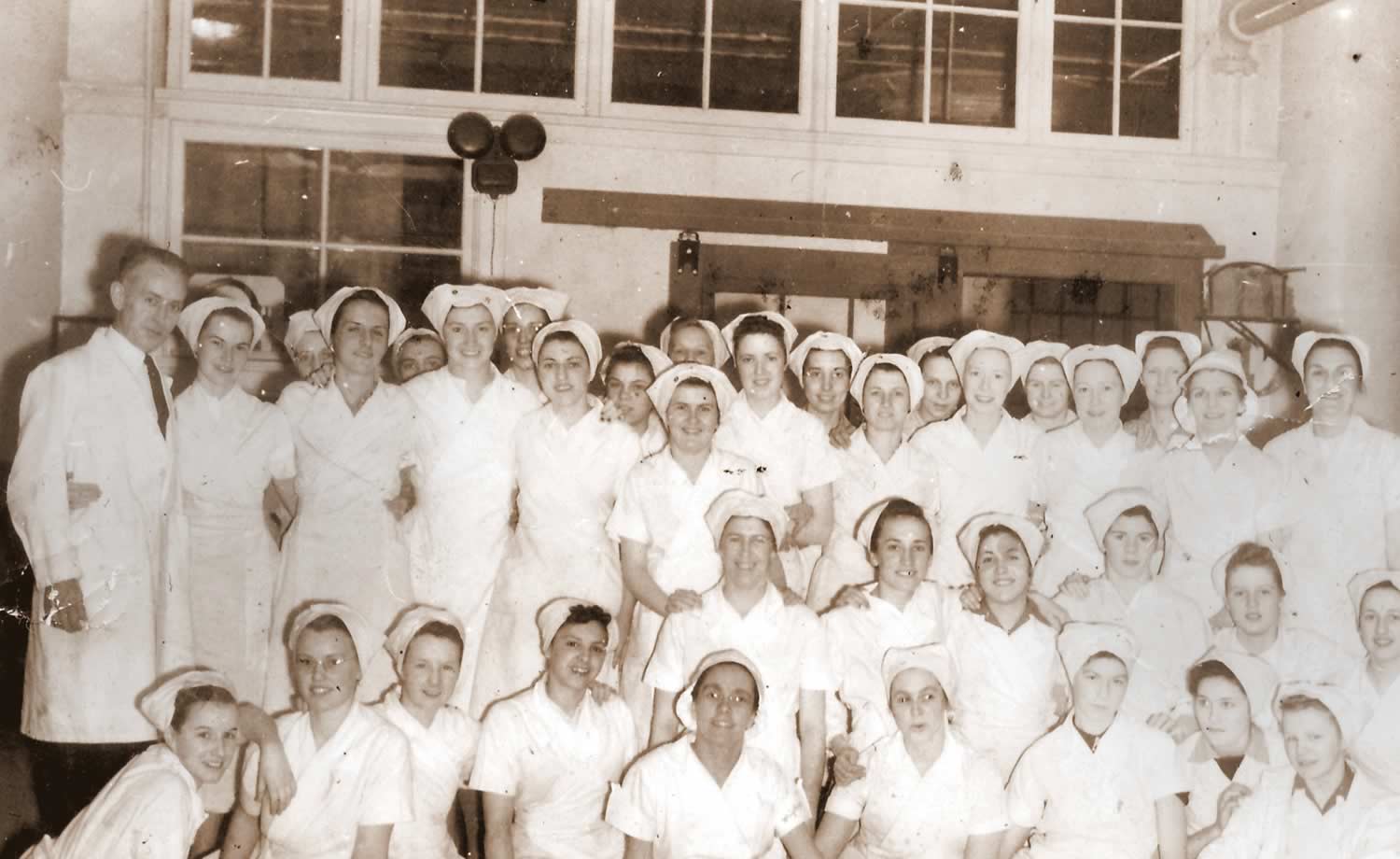 Black and white group photograph of a large group of female factory workers in uniform and one man. All the workers are wearing white jackets and head coverings.