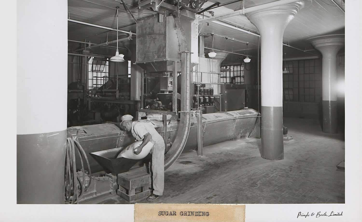Black and white photograph sugar grinding room. Large machine at center of room that goes floor to ceiling. A man is pouring a sack of sugar into the machine. Other large machinery in the background.
