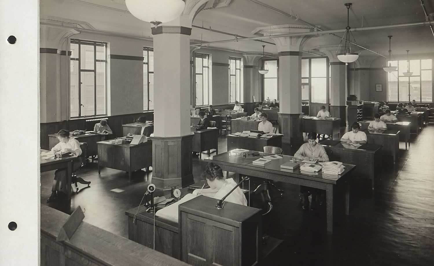 Black and white image. Rows of desks across the photograph, approximately 16 desks total. In the back corner an enclosed office is visible. Mostly women working at the desks. Large pillars in the center of the room, and large windows on the left hand side.
