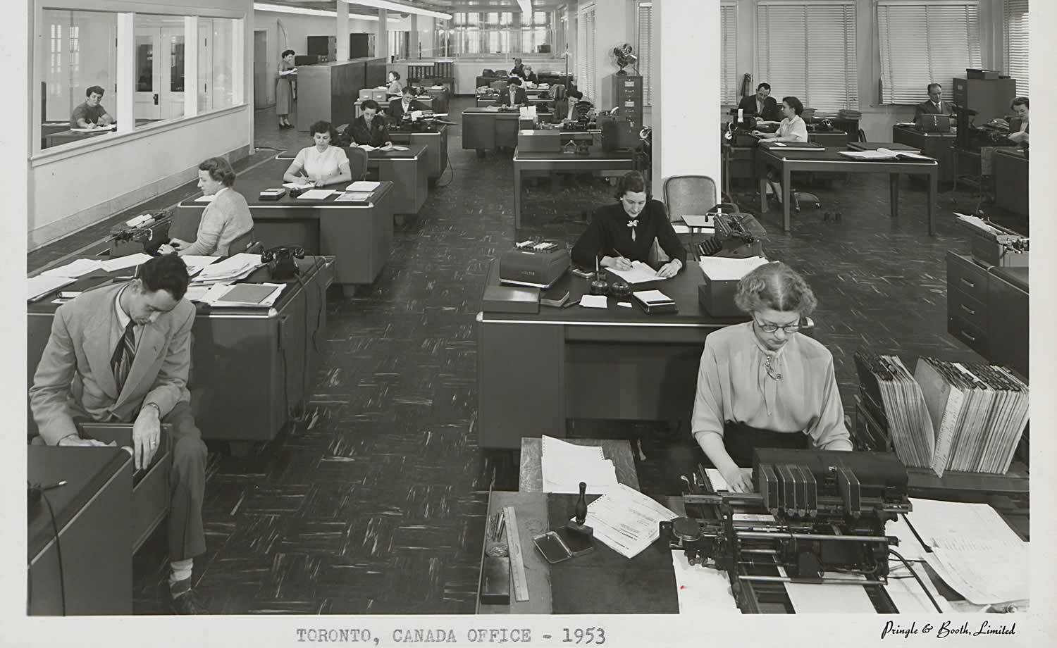 Black and white image of Wrigley Factory office space. Large room with rows of desks. Mostly women at the desks. Women at the front is typing on a typewriter, and closest man is filing paper into his desk.