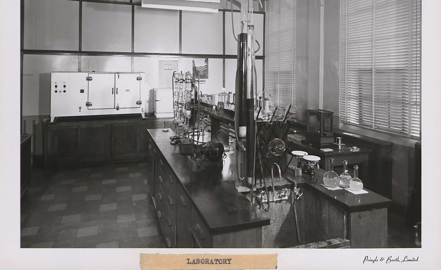 Black and white photograph of a laboratory at Wrigley Building. In the center of the room is a desk with science equipment(beakers, measuring cups, sink). The other walls also have larger related equipment.