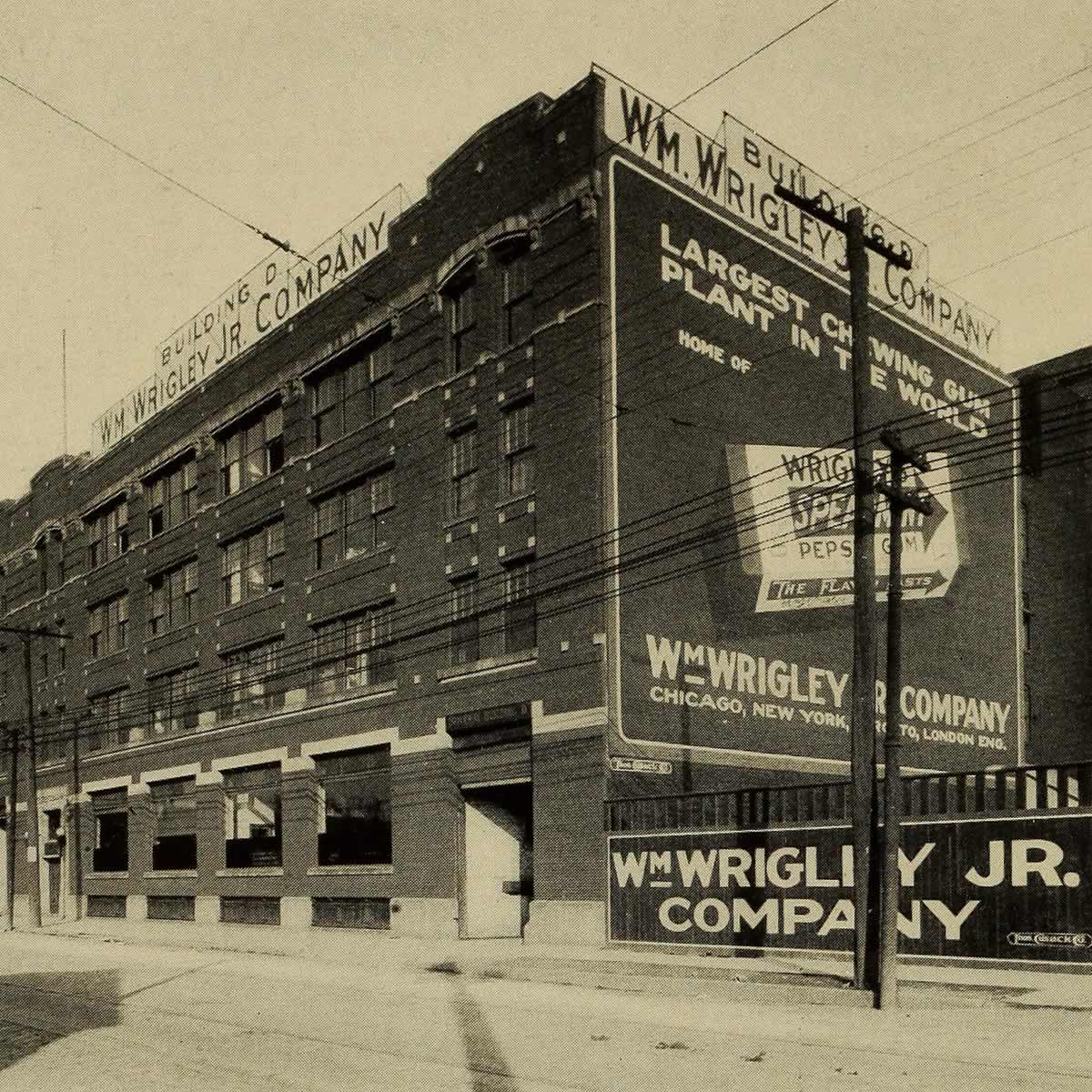 Black and white photograph of a large brick industrial building with a large advertisement for Wrigley chewing gum on one of its walls.