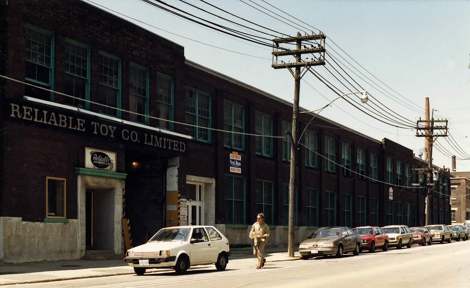 Colour photograph of the Reliable Toy factory on Carlaw Avenue. The factory is closed and there are 'For Rent' signs on the outside of the building. Several cars are parked outside and a delivery person is walking with a package.