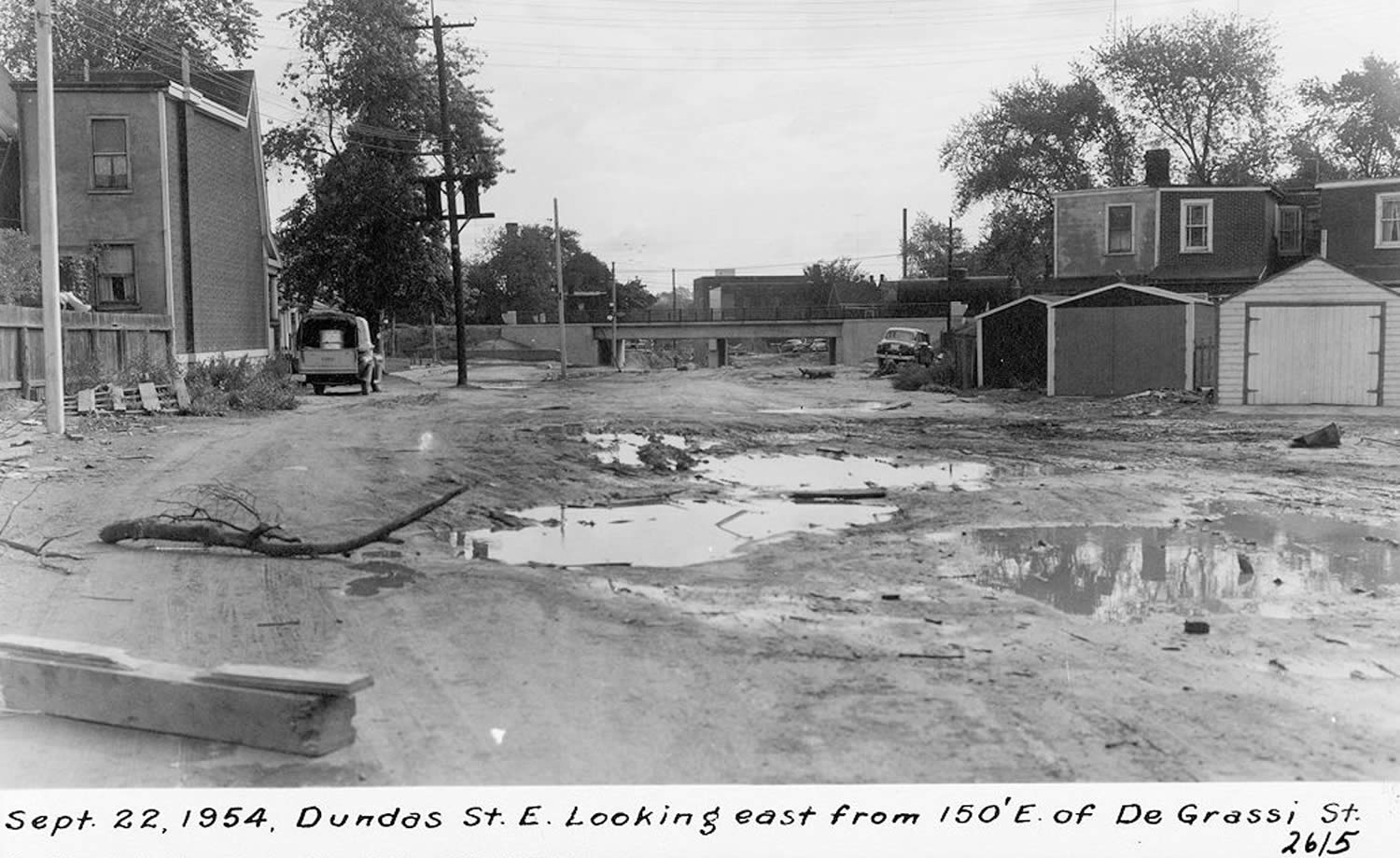 Black and white photograph of an unpaved, muddy street with a rail overpass in the background. Houses are on both sides of the street, with their garages visible.