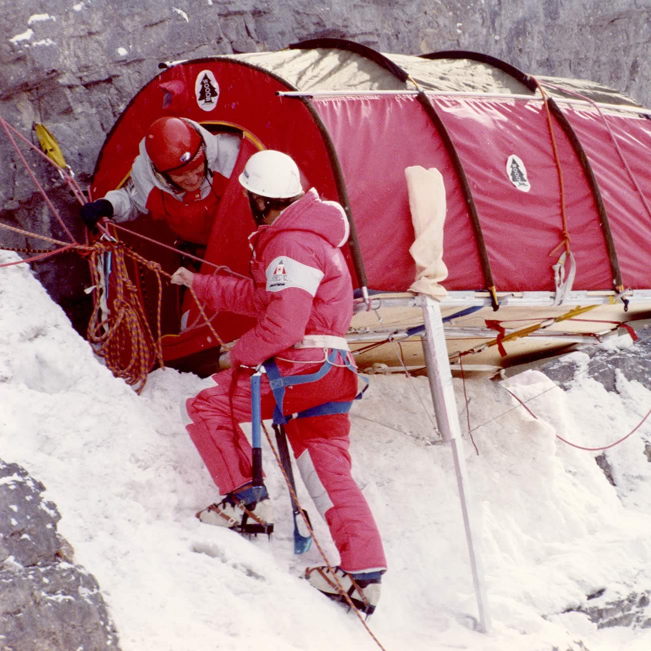 A tubular red Woods tent with ropes and metal legs hangs from the vertical rock face of a mountainside. A climber emerges from the door while another is outside, tethered with a harness and ropes.