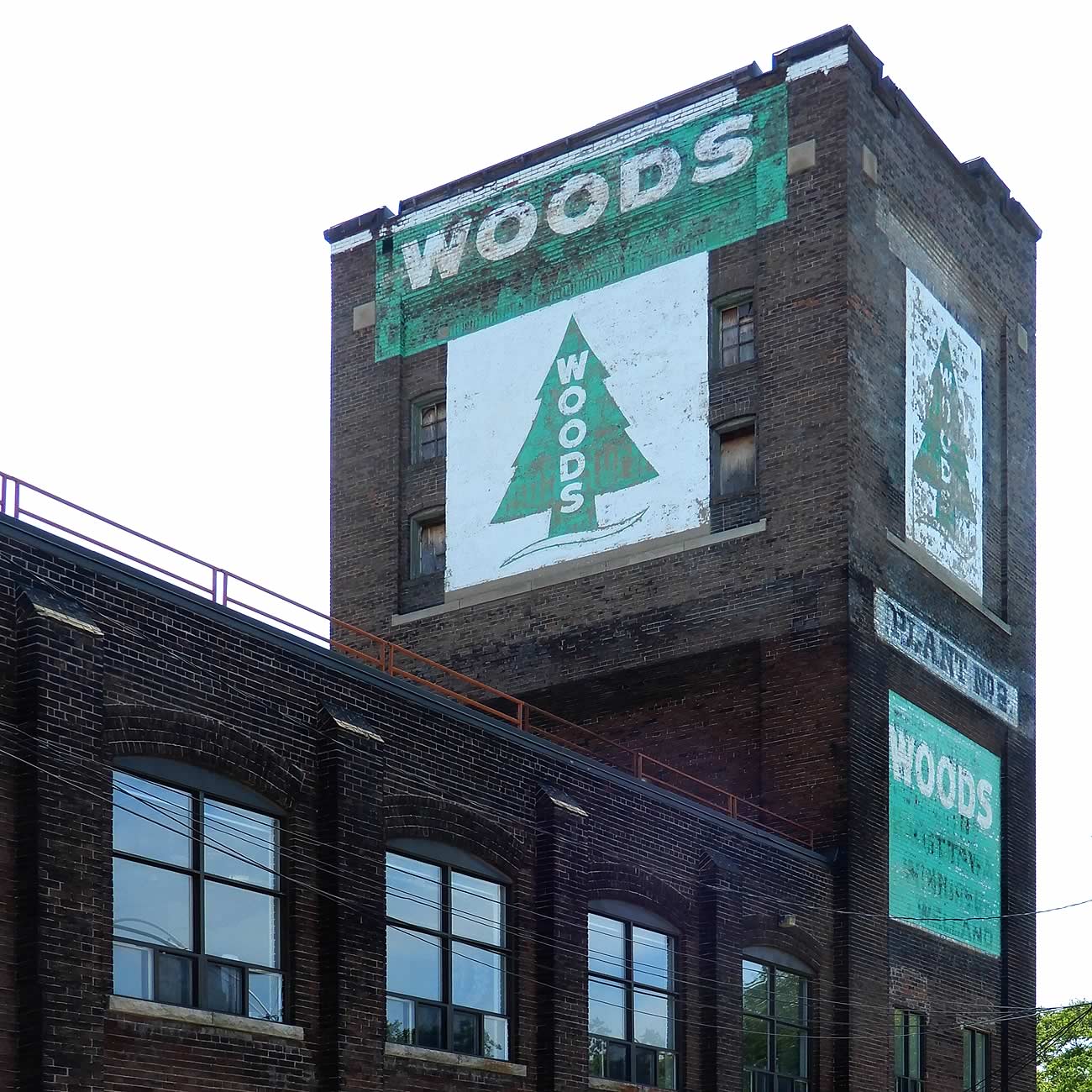 Colour image of an industrial brick building with a water tower at the end. The water tower is painted with a logo for 'Woods': an evergreen tree with the words Woods vertically across.