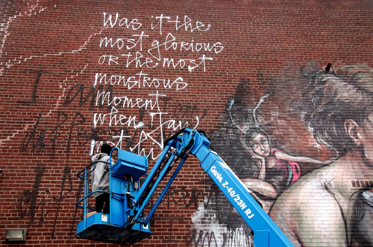 Colour image of a person in a lift painting a large mural in a brick wall. The person is working on the top corner, writing the sentence 'Was it the most glorious or the most monstrous moment when Jay met his creative spirit?'