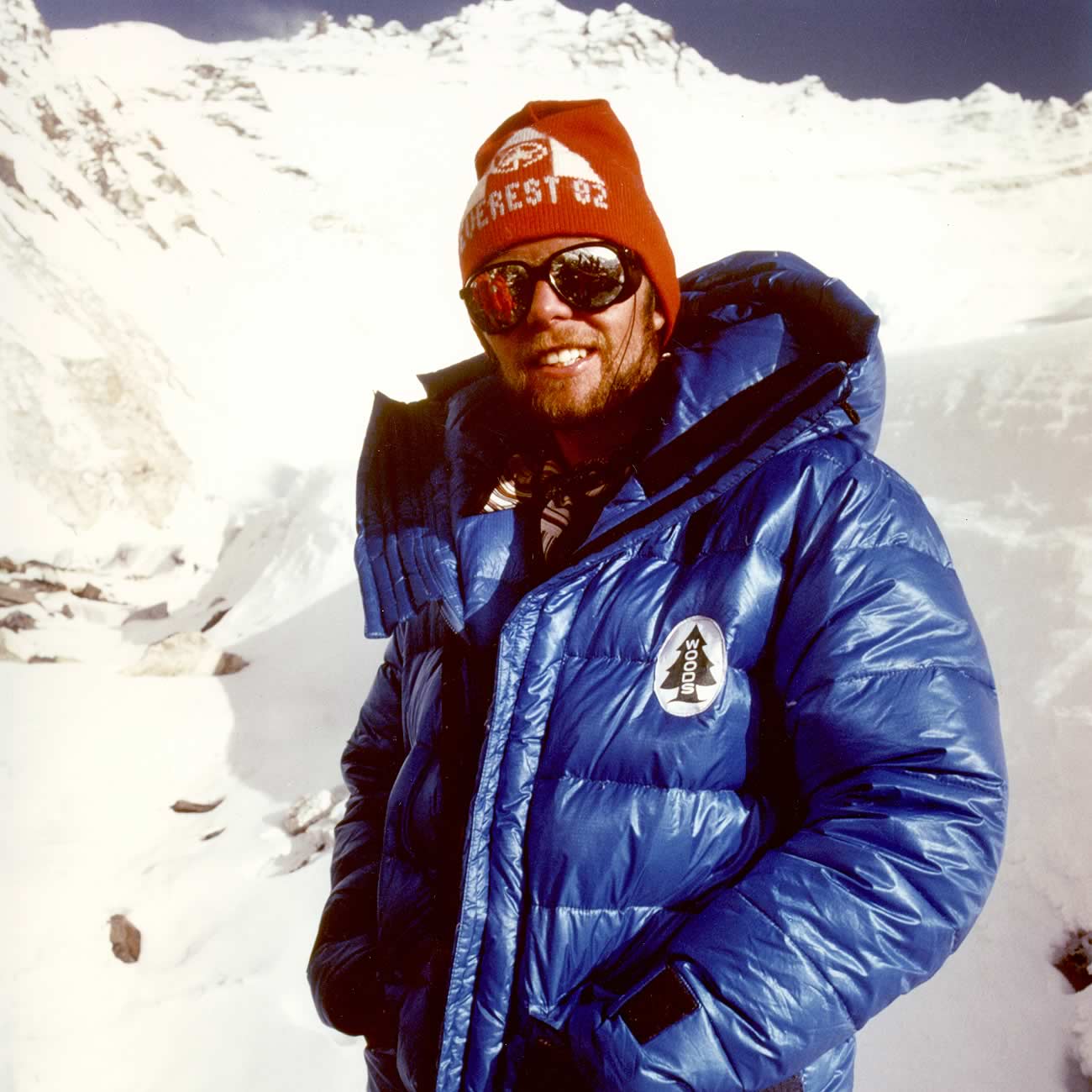A young male mountaineer wearing reflective sunglasses, a red Everest '82 expedition hat, and a thick blue Woods jacket on a snowy mountainside.