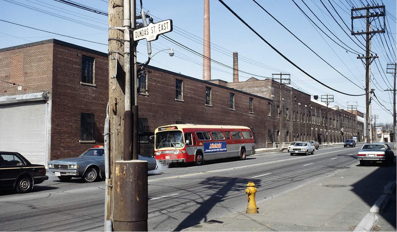 Colour image of Carlaw Avenue at Dundas Street East. A red, cream, and silver TTC bus is in the foreground heading south.