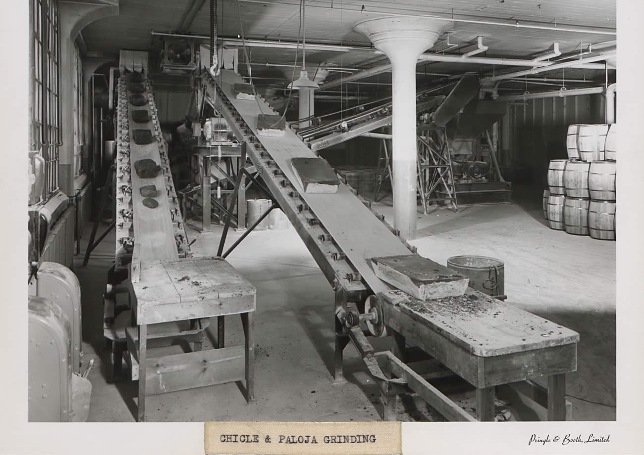 Black and white photograph of grinding room. At the forefront are two conveyor belts that lead to floor above; there is something on both conveyor belts. A third conveyor belt is in the background as well. In the right corner is a stack of barrels.