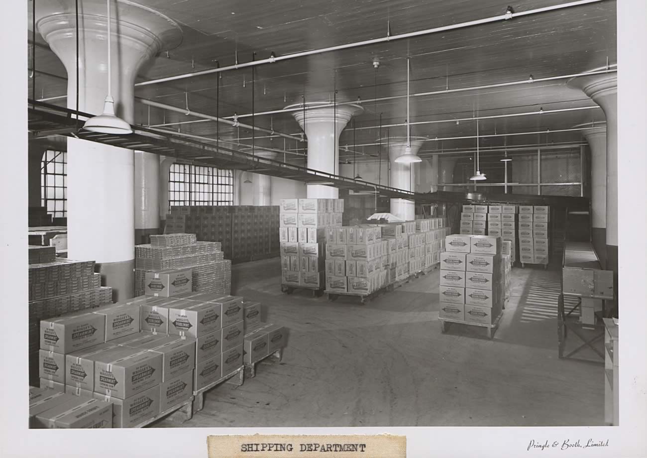 Black and white photograph of shipping department; it is a long room filled with pallets of product to be taken out. Pallets have up to 6 boxes stacked on each other. There is a conveyor belt coming from the floor above that sends boxes down to be shipped.