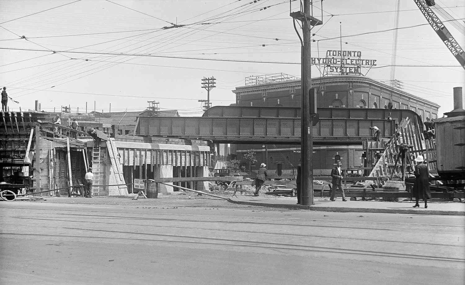 Black and white image of Carlaw Avenue from Gerrard Street East. Workers are building a rail bridge and a Toronto Hydro-Electric System substation is in the background.