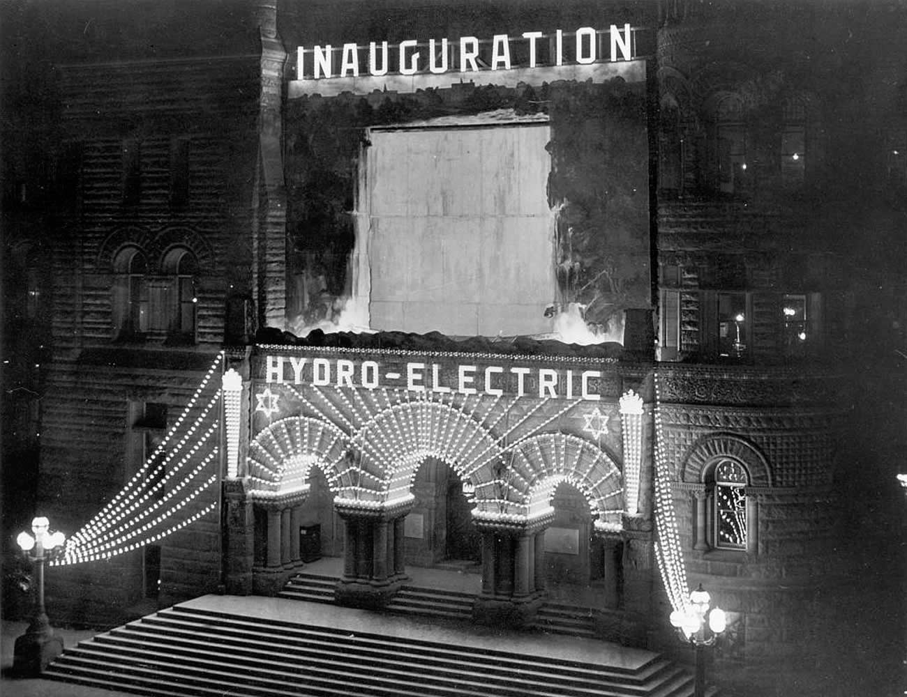 Photograph shows the front of Toronto City Hall at night. The facade is lit up with electric lights reading 'Inauguration, Hydro-Electric.' Above the main doors of City Hall is a picture of Niagara Falls to show where the hydro-electric power originated.